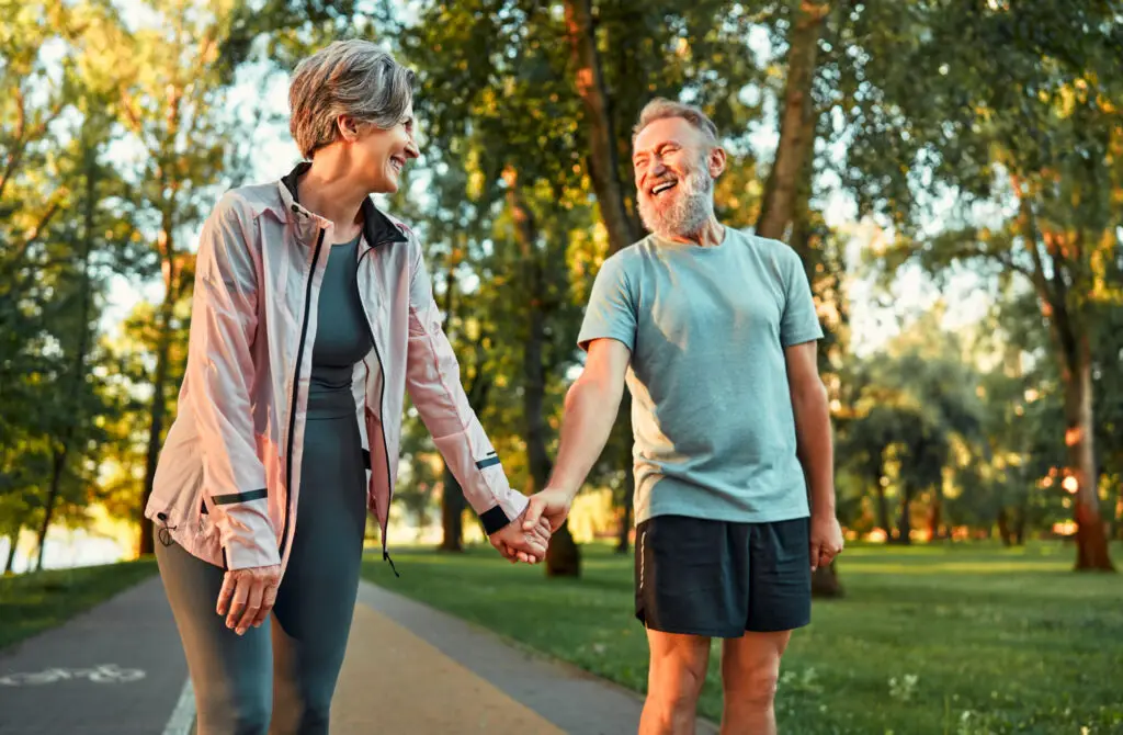 Cheerful modern senior couple outdoors in the park dressed in sportswear walking holding hands and laughing. Morning sun rays fall on smiling man's face.